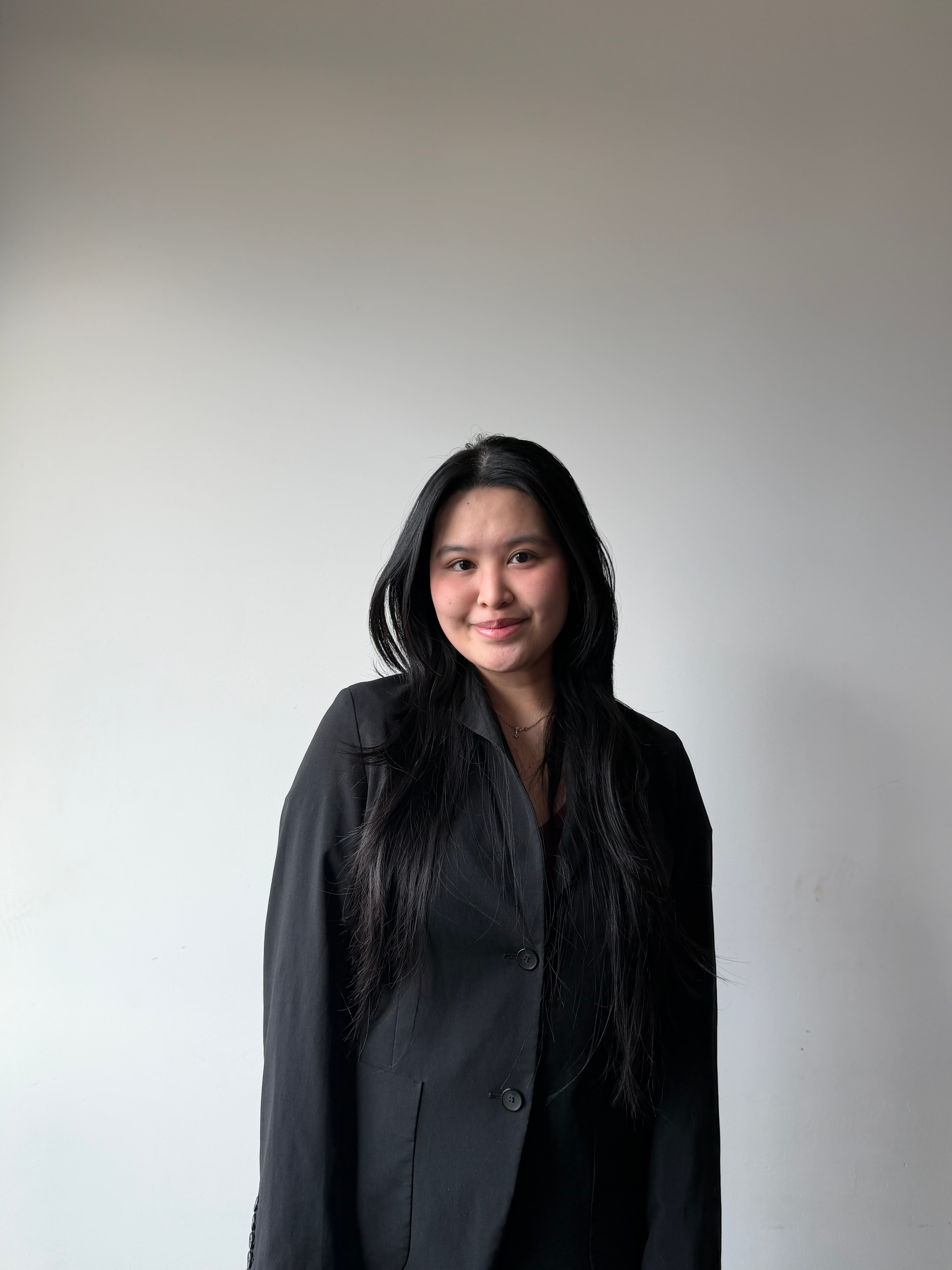 Smiling young woman with long black hair wearing a black blazer against a white wall.