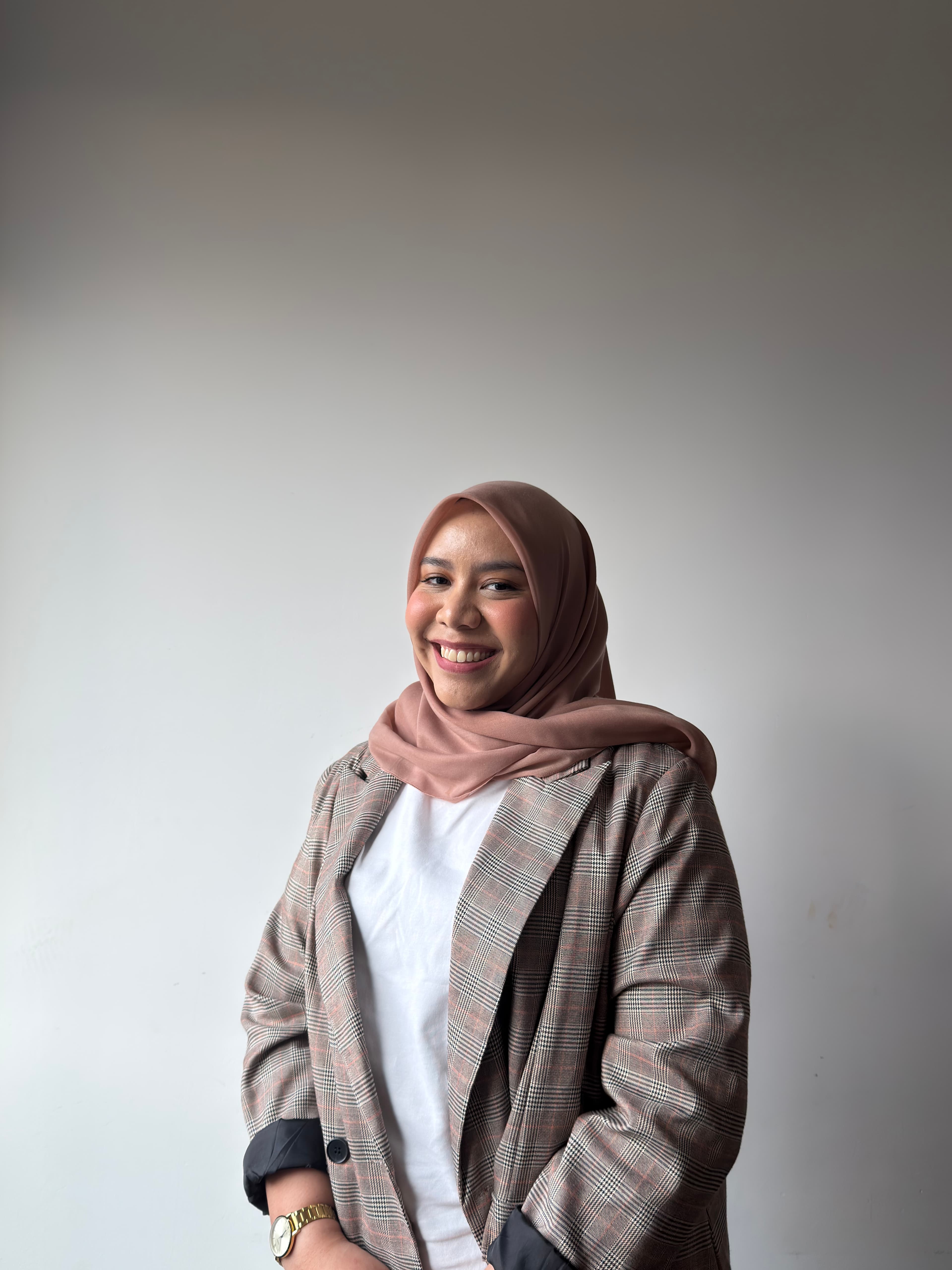 Smiling woman wearing a pink hijab and a plaid blazer against a plain background.