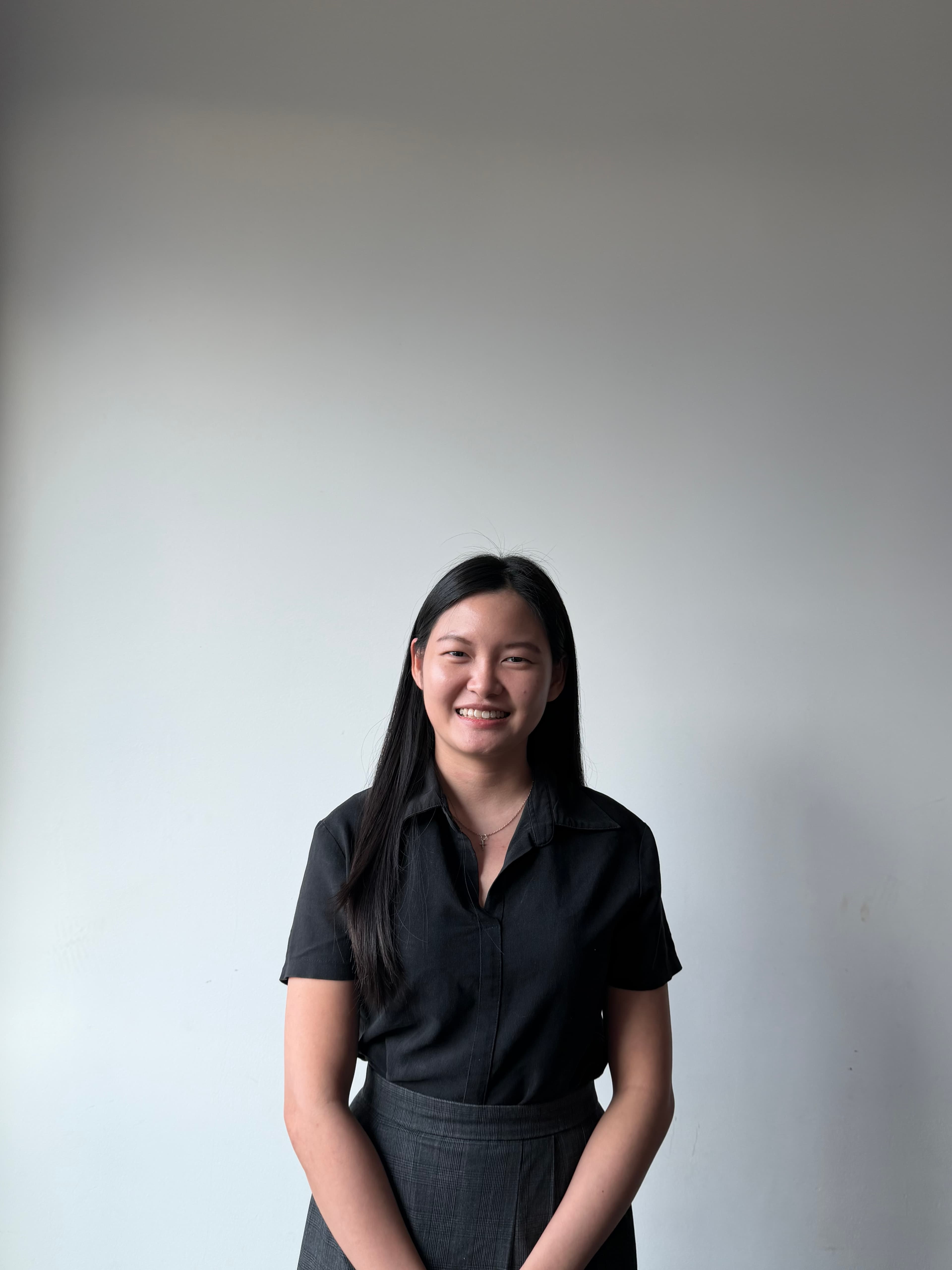 Young woman with long black hair smiling, wearing a black collared shirt and grey plaid skirt.